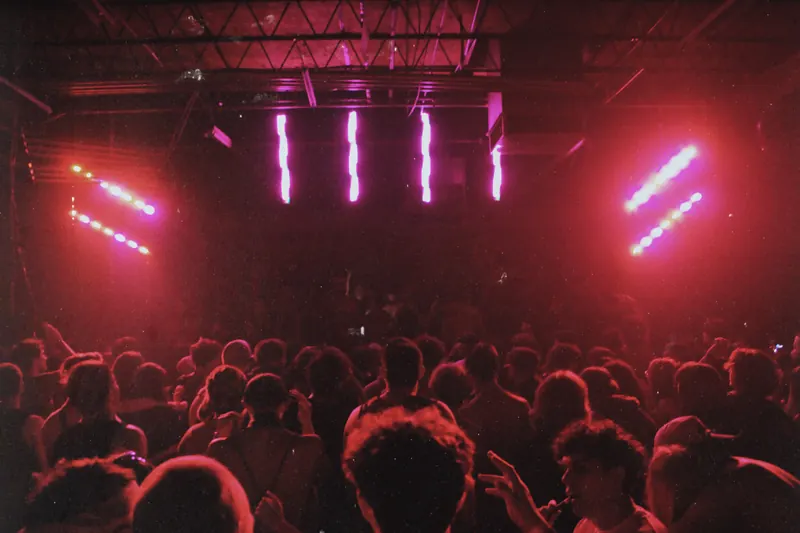 Red and pink lights illuminate the warehouse ceiling and crowd.