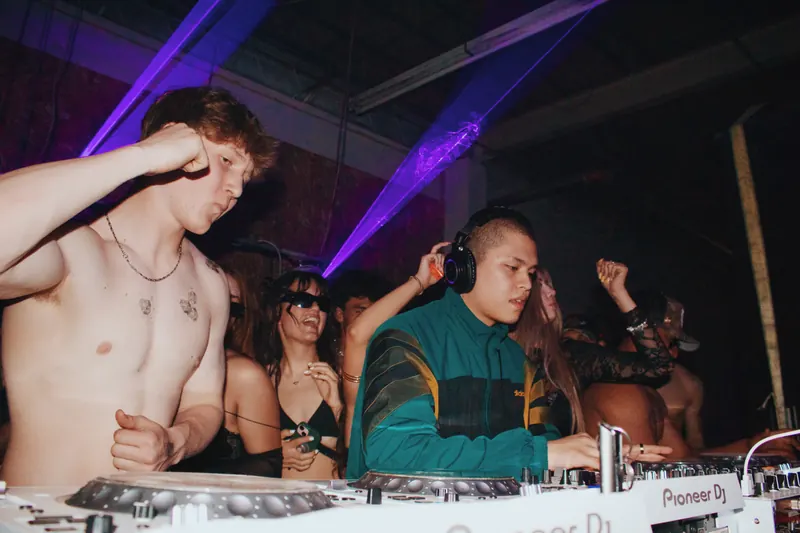 Two DJs at the decks with guests gathered behind them.
