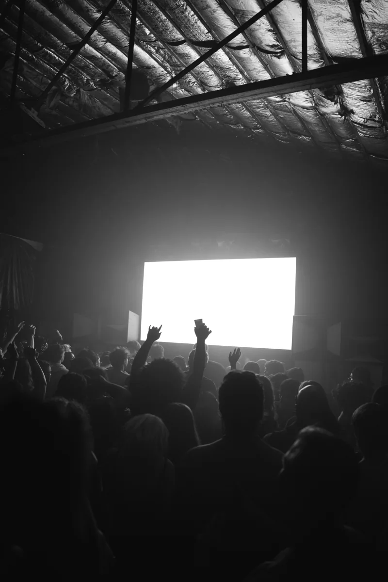 A black-and-white crowd with raised hands beneath the warehouse roof.